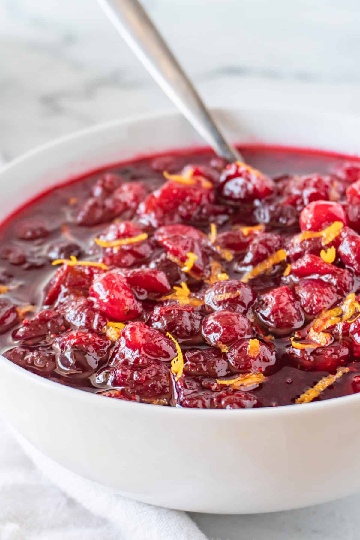 Cranberry sauce in a white bowl with a spoon sticking out and orange zest as a garnish.