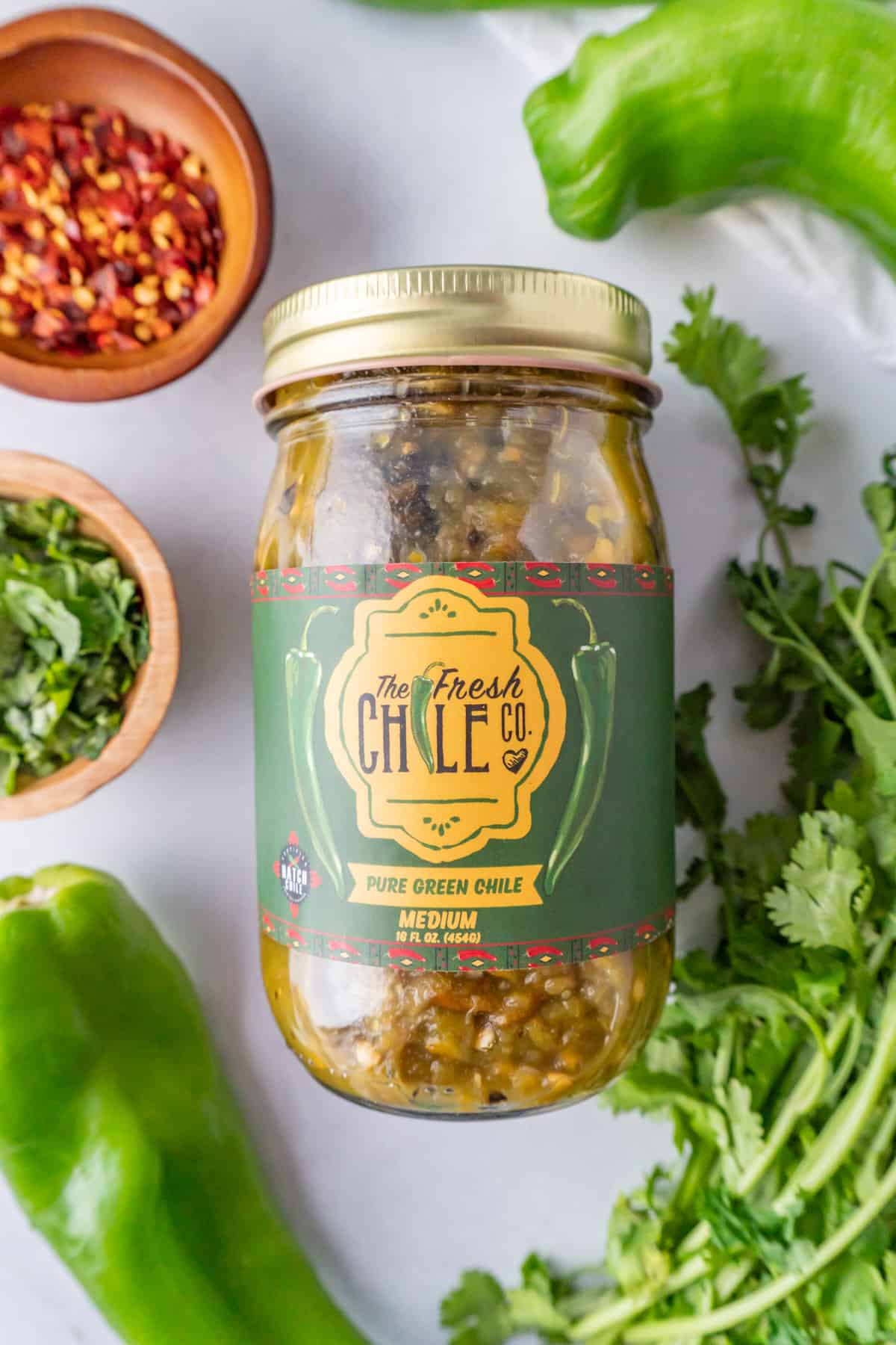 green chile jar with cilantro, green chile peper, and red pepper flakes around it