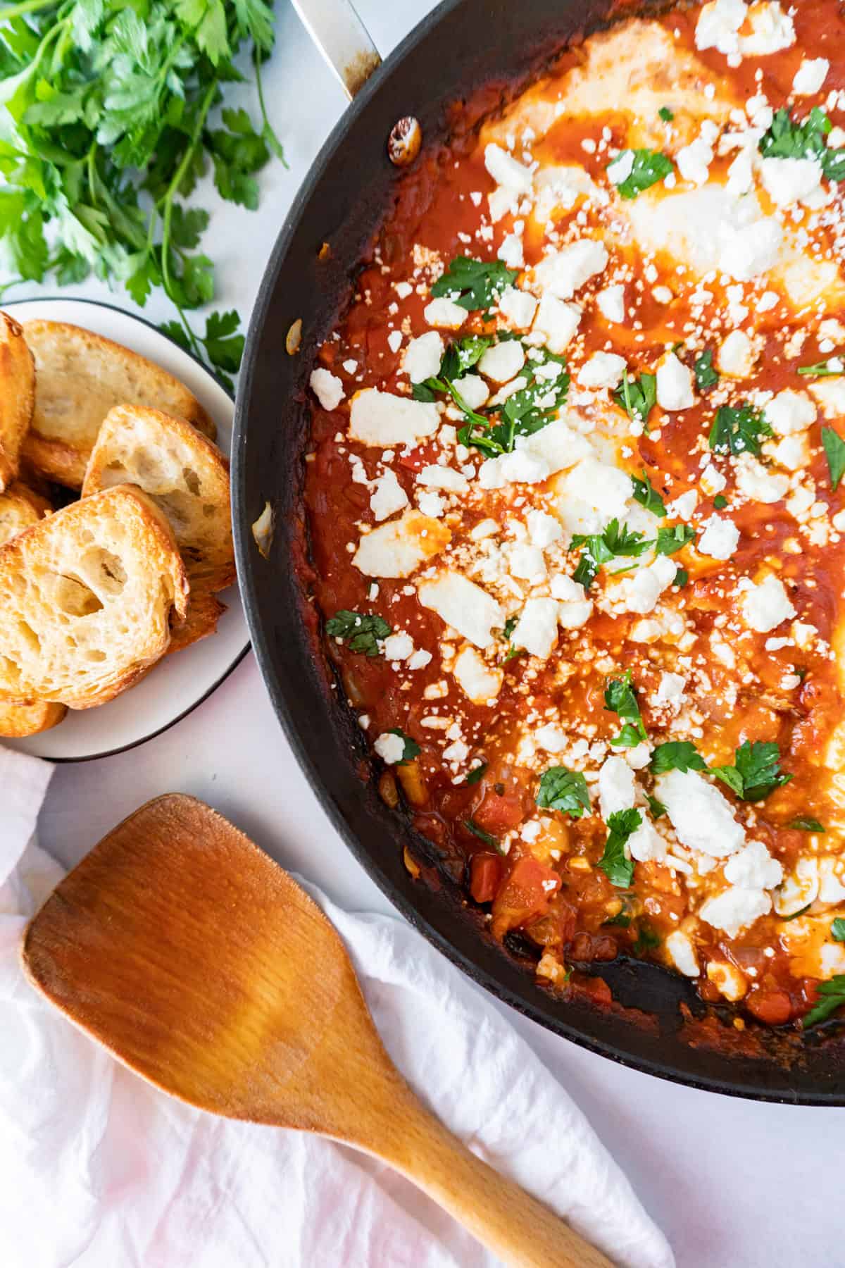 shakshuka in skillet, wooden spoon, toasted bread, parsley