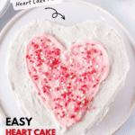 Heart cake on white cake plate with small bowl of sprinkles next to it.