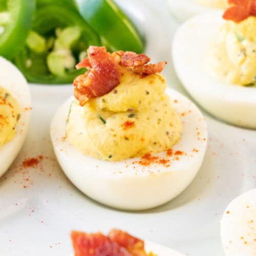 jalapeno deviled egg with bacon garnish on white platter with fresh jalapeno slices in background.