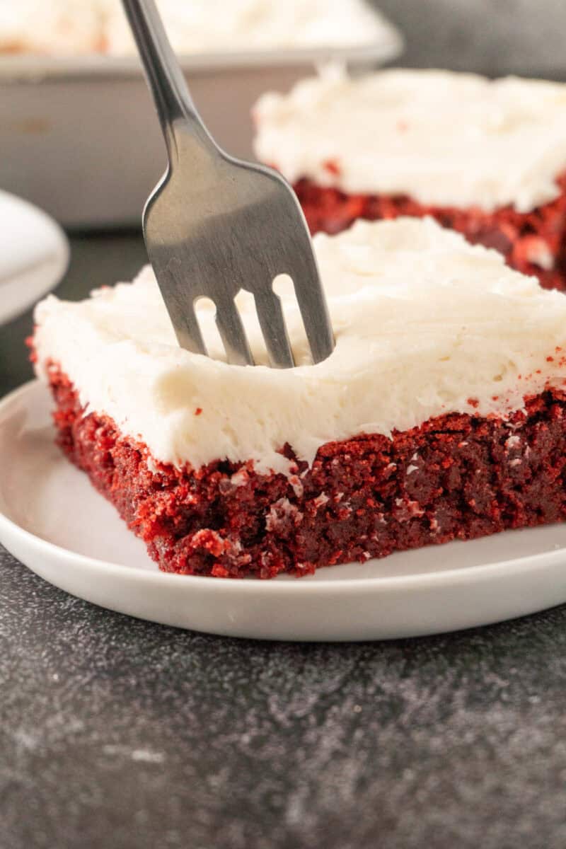 sticking a fork in a frosted piece of red velvet brown sitting on a white plate.