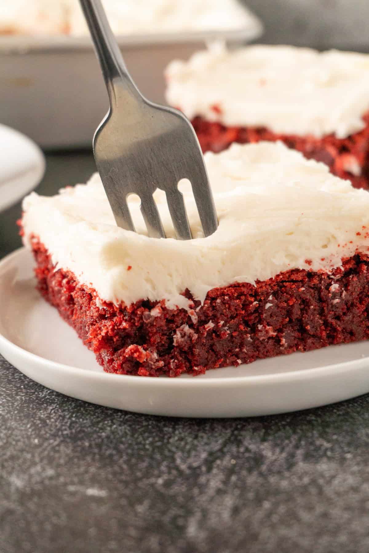 sticking a fork in a frosted piece of red velvet brown sitting on a white plate.