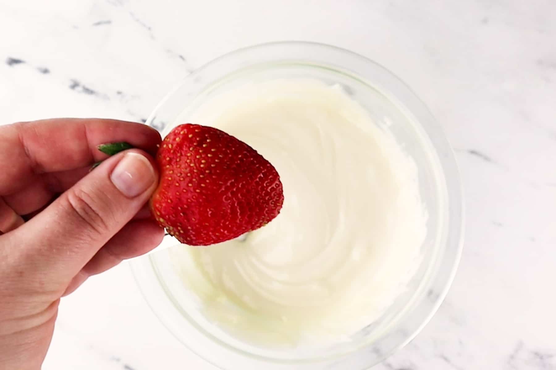 holding strawberry by green leaves at top over bowl with melted white chocolate in it.