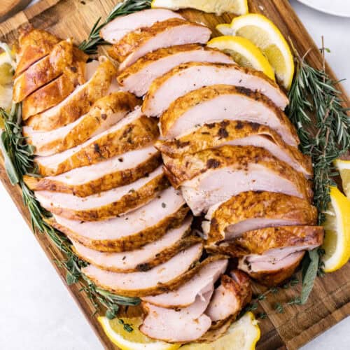 smoked turkey breast sliced on a wooden cutting board, surrounded by rosemary and lemon slices.