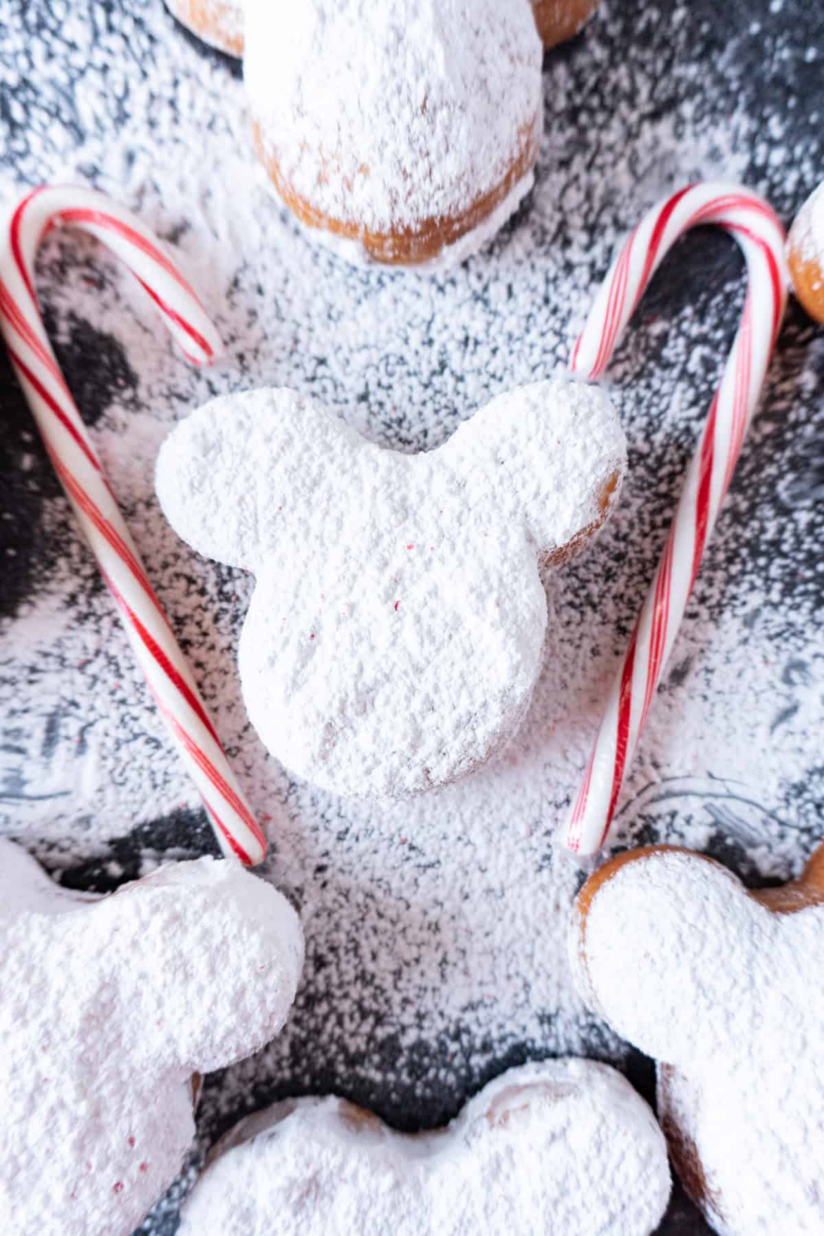 candy cane beignets on dark background covered in powdered sugar and next to candy canes.