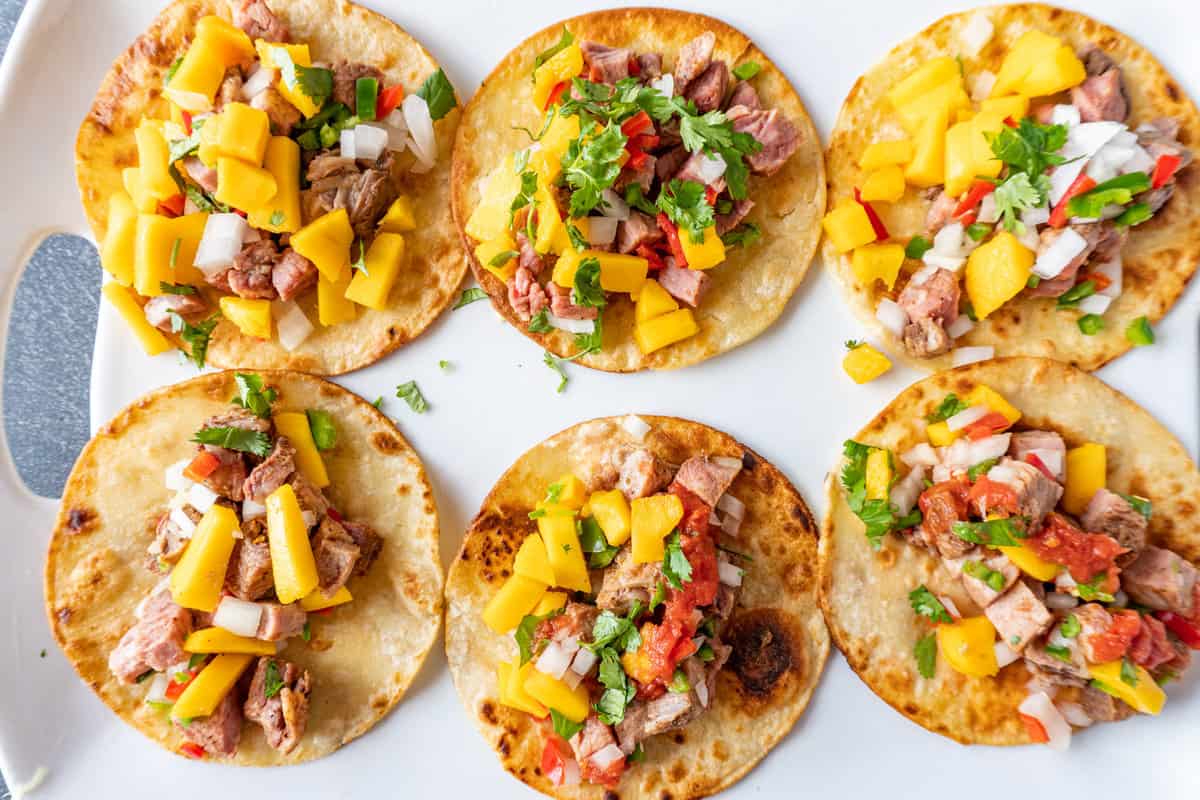 Six tri-tip tacos on a white platter. All tacos have cilantro, onion, and mango on top.