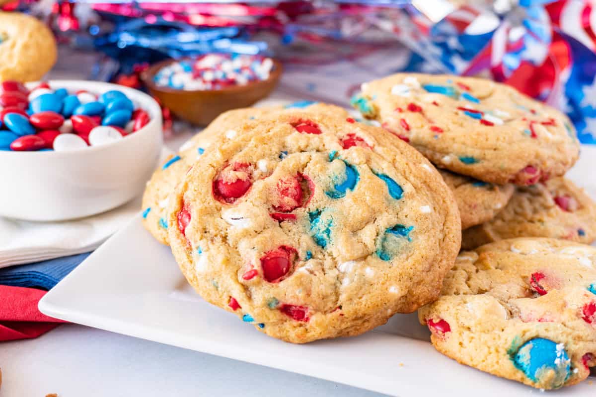 cookies with red, white, and blue candies baked into them on a white plate, with 4th of July decorations in the background.