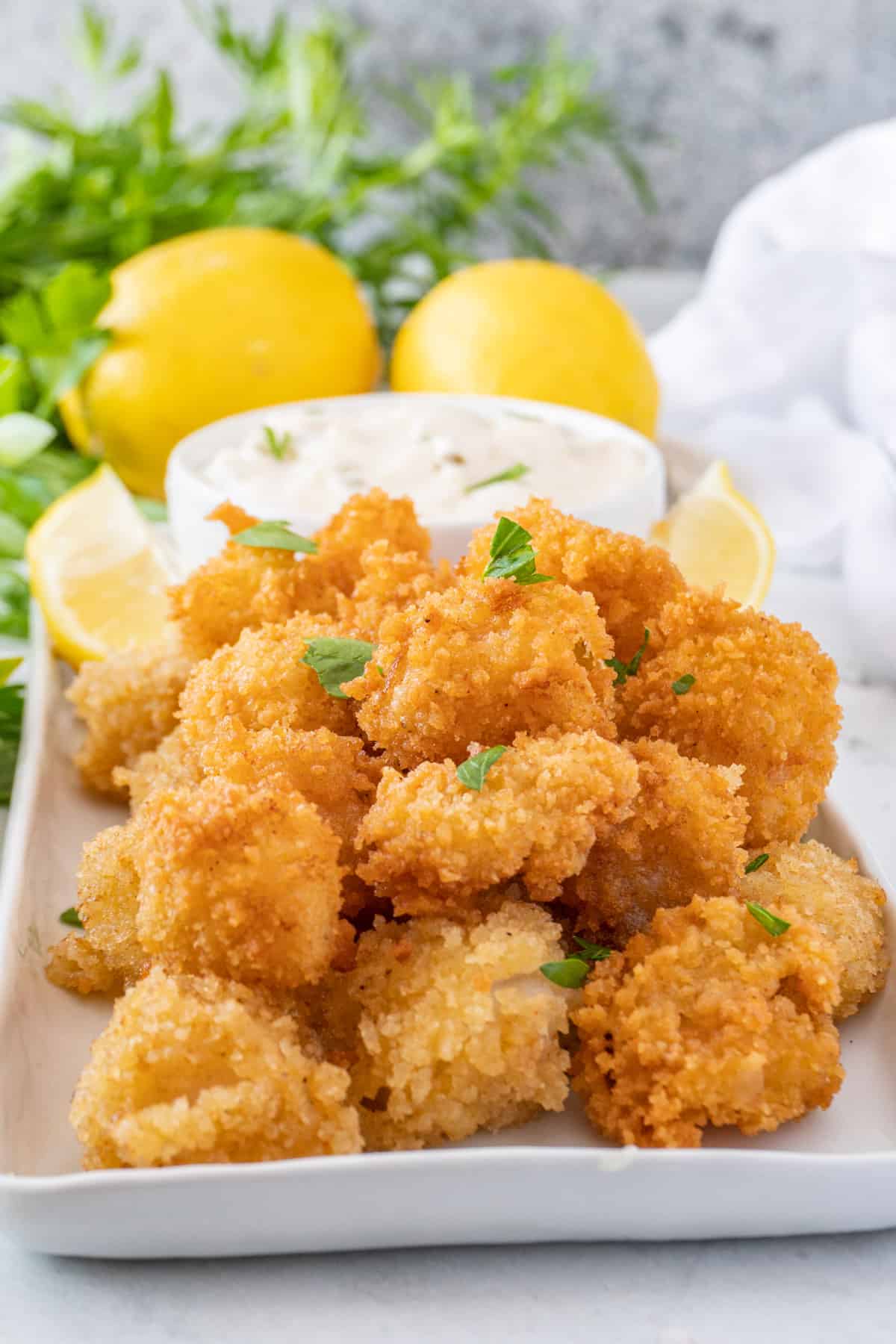 fried scallops on a white plate garnished with chopped parsley, bowl with tartar sauce, and sliced lemons near by.
