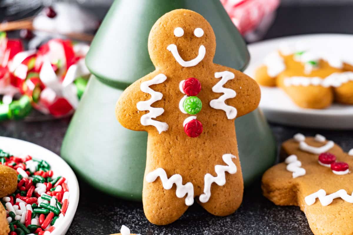 gingerbread man cookie propped upright against green tree figurine. Cookie is decorate with royal icing and sprinkles. More cookies in background.