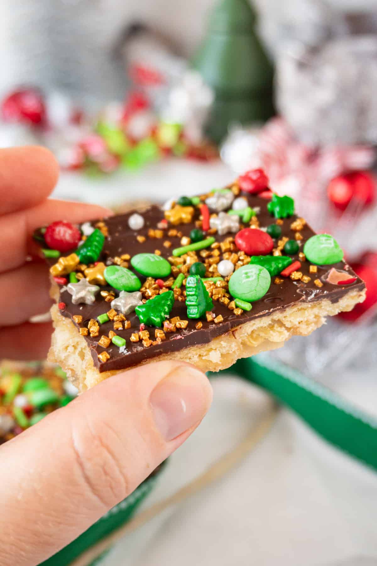 holding a piece of Christmas Crack with Christmas Decorations in background.