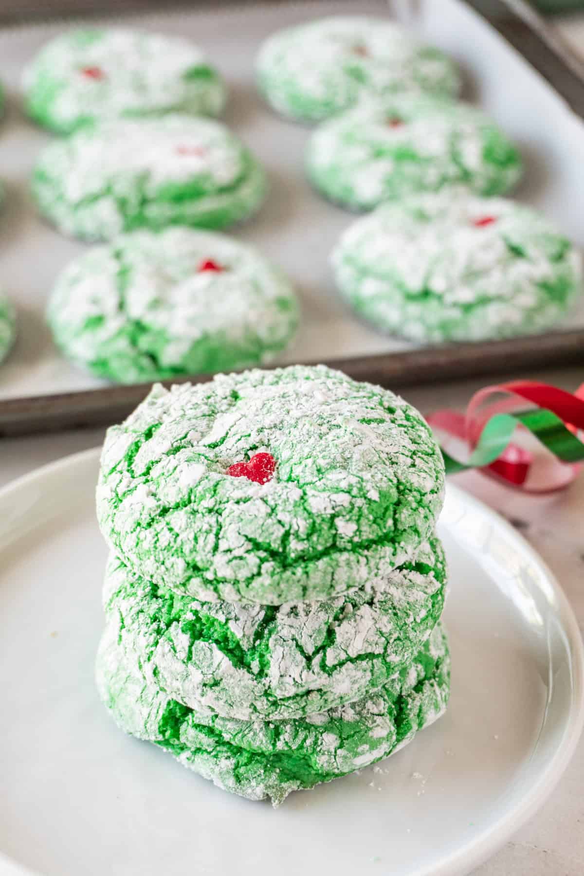 Grinch Cookies stacked 3 high on small white plate with more cookies in the background.