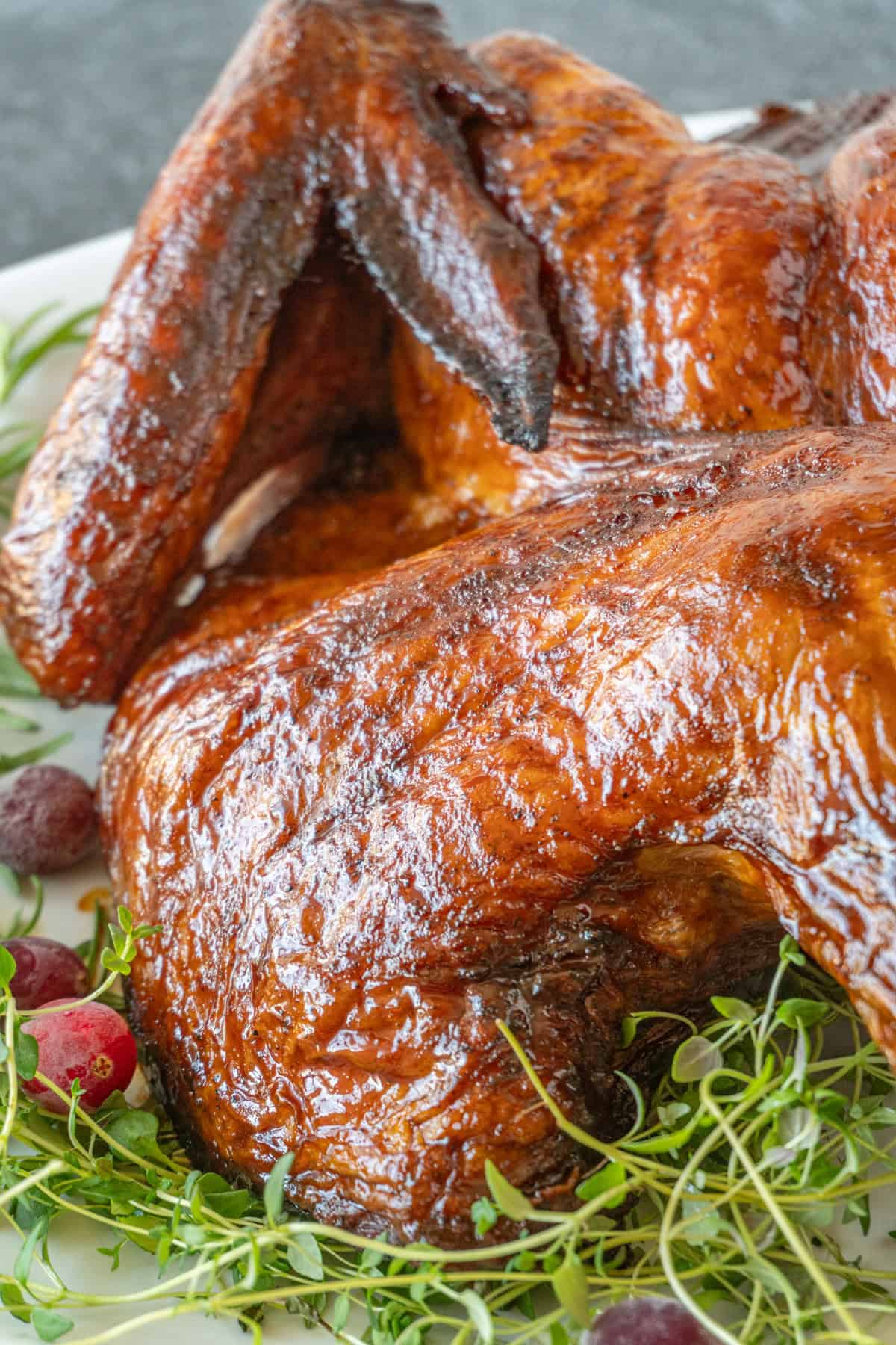 smoked turkey with browned skin on a white platter surrounded by herbs and cranberries.
