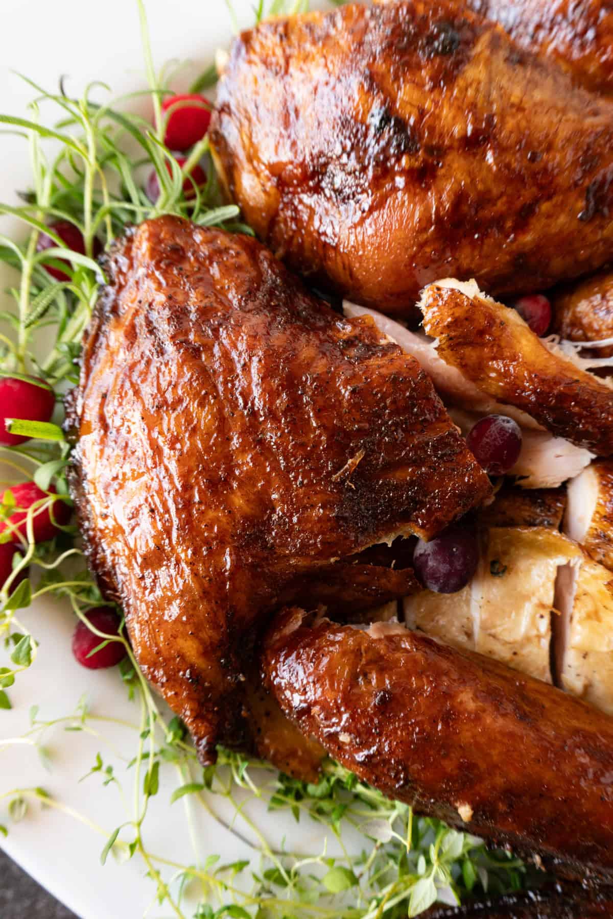 carved turkey thigh with deep reddish brown skin on white platter surrounded by herbs and cranberries.