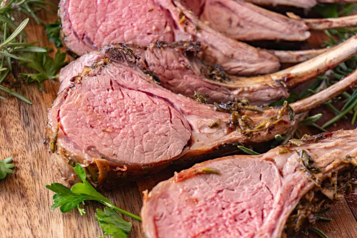Herb Crusted Rack of Lamb sliced. on a cutting board with fresh herbs next to it.