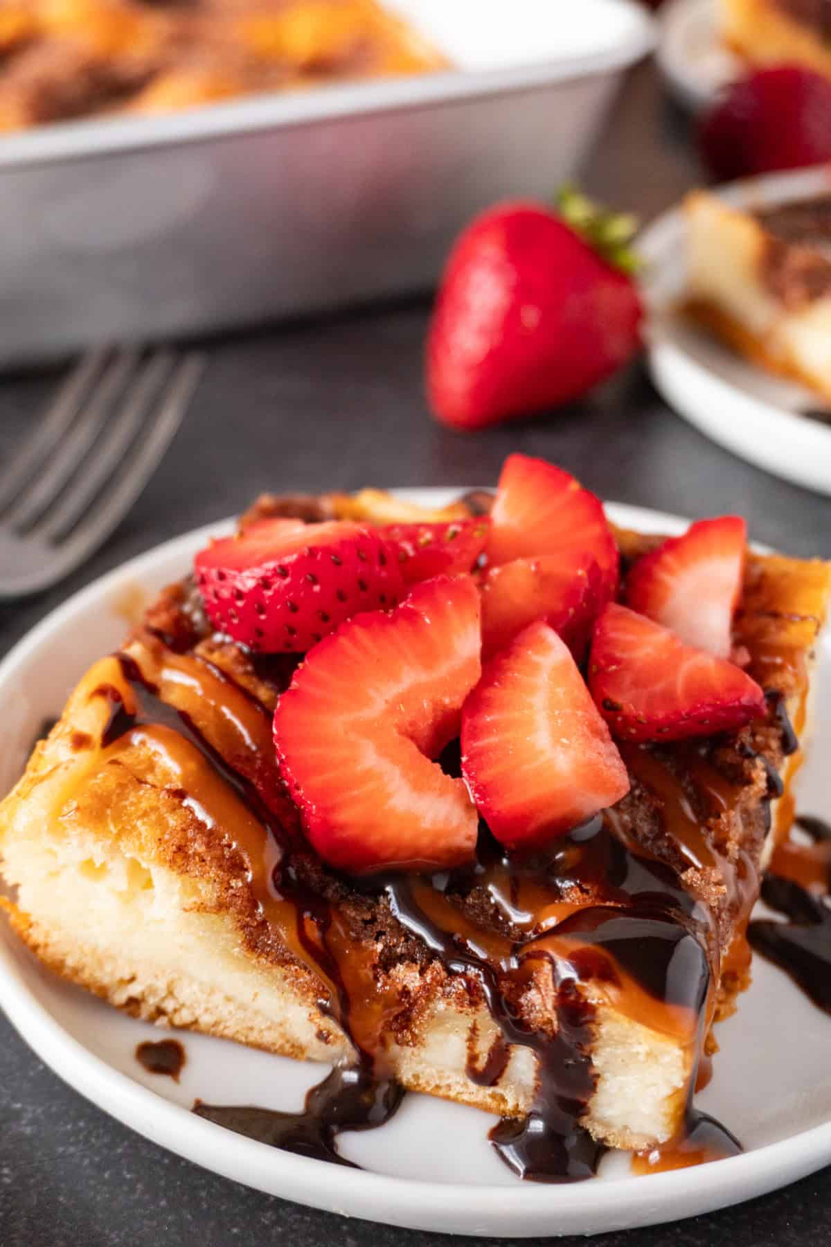 churro cheesecake bar topped with caramel sauce and chocolate sauce and strawberries on a white plate. next to a fork.