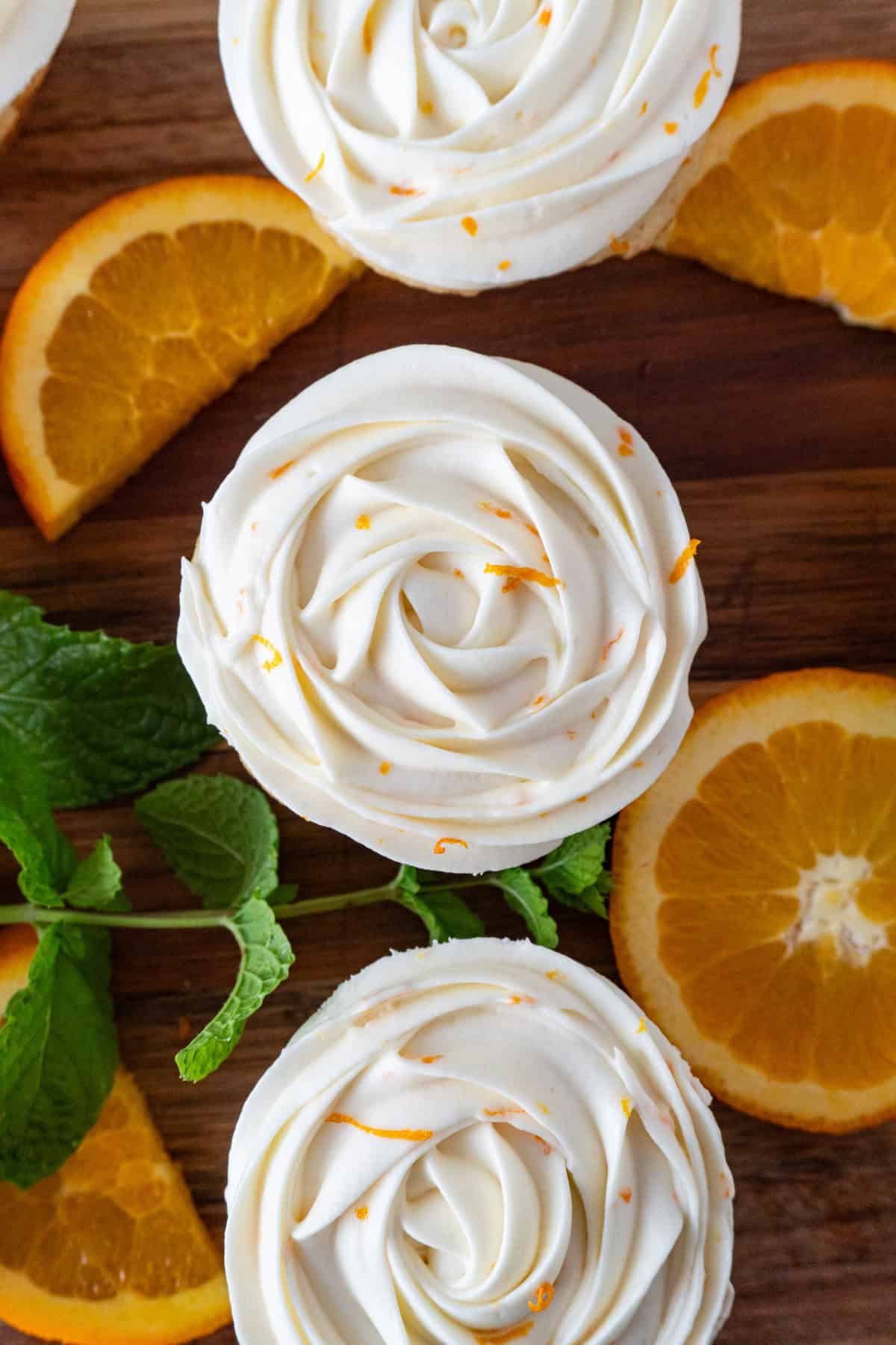 Orange cupcakes with cream cheese frosting on a cutting board surrounded by orange slices and mint leaves.