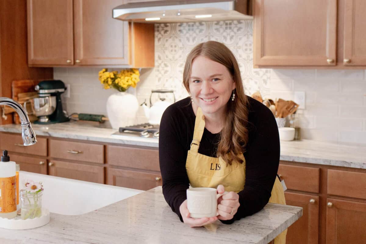 Lisa in kitchen wearing yellow apron and black long sleeve, holding a coffee mug.